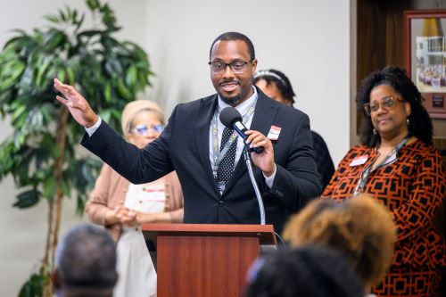 A head-on shot of a gentleman speaking into a microphone at a podium, wearing a blazer, button-down shirt, and tie. He wears glasses, and is looking slightly to his left while gesturing with his left hand.